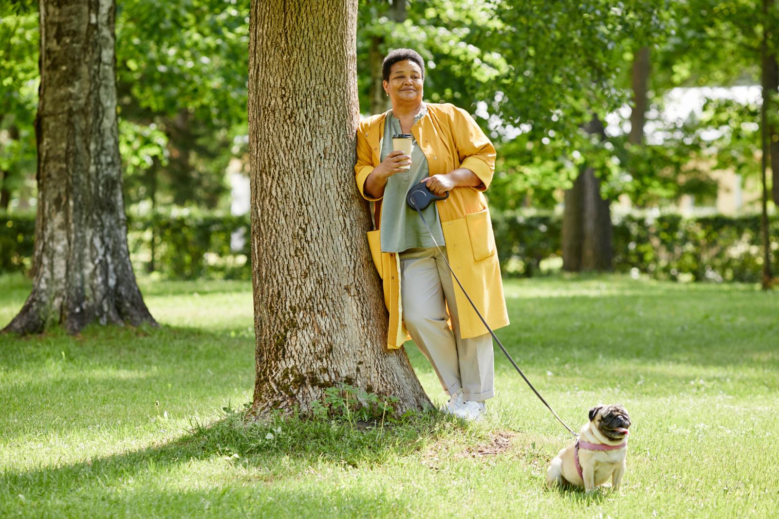 Full length portrait of senior black woman leaning on tree in park while walking cute dog and drinking coffee, copy space.