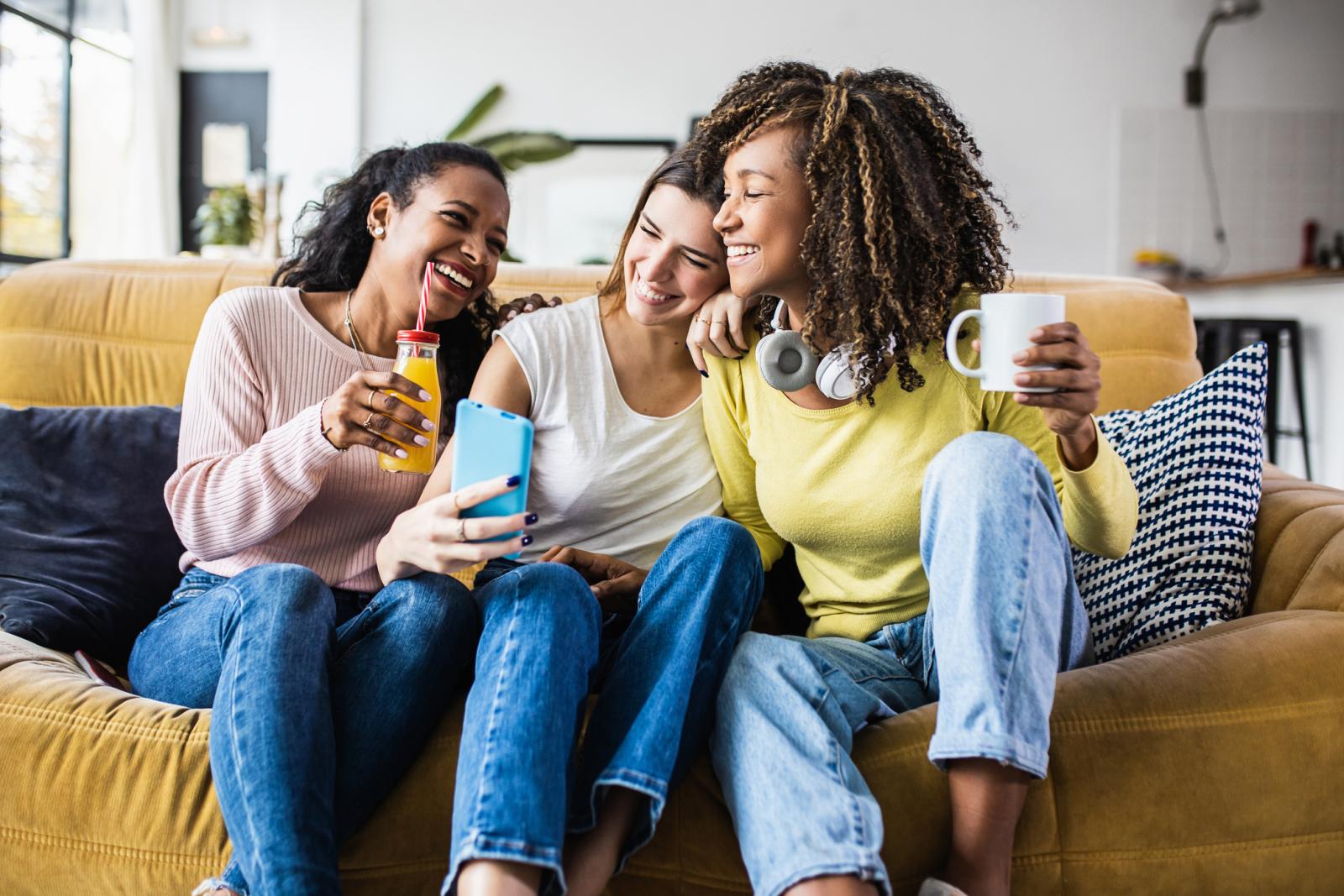 Three young women sitting on a mustard-yellow couch laughing and taking a selfie together, enjoying drinks in a bright and modern apartment living room.
