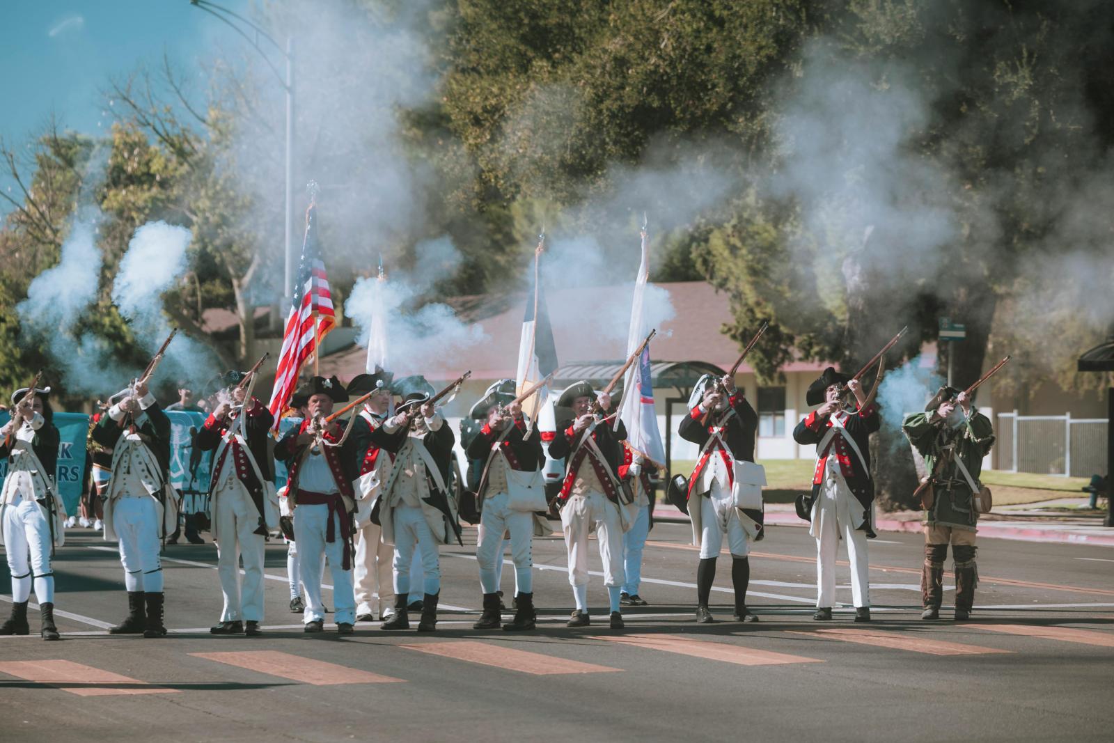 Historical reenactors in Revolutionary War-era uniforms fire muskets during a parade in York County, Pennsylvania, with smoke in the air and American flags held high